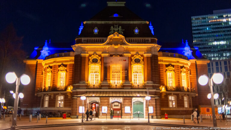 Illuminated Laeiszhalle at night, glowing amber and blue with symmetrical windows and globe streetlamps in front.