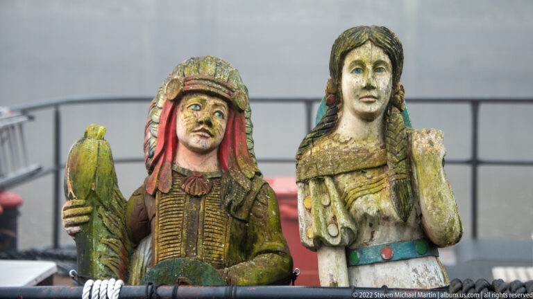 Two weathered painted wooden figures, one with a headdress and green object, the other in a pale garment.