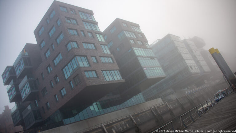 Angular brick-and-glass waterfront buildings in Hamburg rise into heavy fog above a railing-lined promenade.