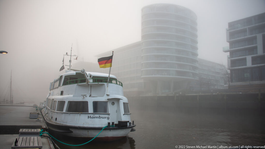 White harbor passenger vessel labeled “Hamburg” moored at a floating dock in dense fog beneath a German flag.