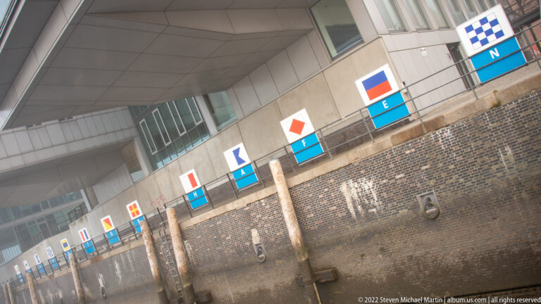 Row of lettered maritime signal-flag panels mounted along a brick quay wall with timber fenders and mooring rings.