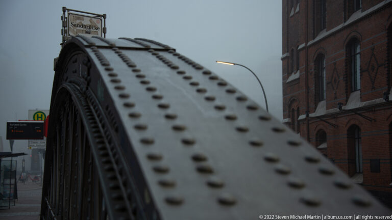 Close view of a riveted steel bridge arch with dark latticework, brick building, and transit sign behind.