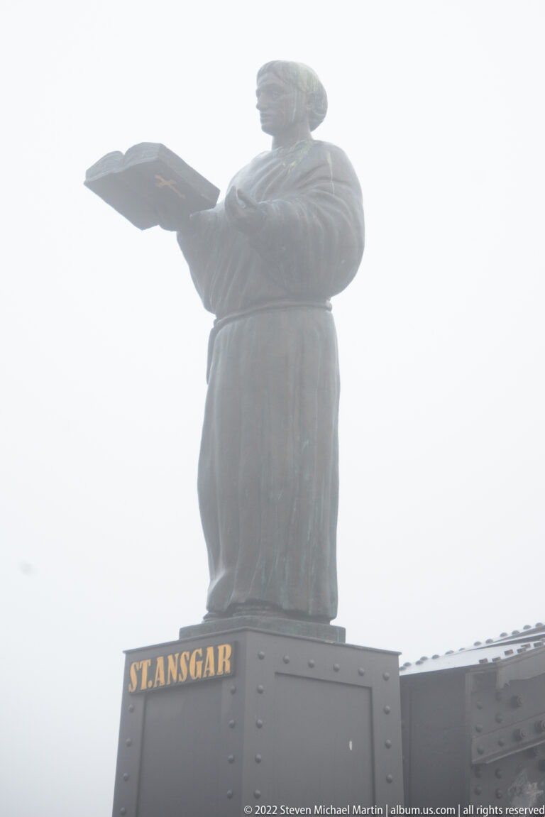 Robed statue of St. Ansgar holding a broad book above a bridge pedestal labeled “ST. ANSGAR.”