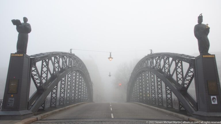 Fog-shrouded Brooksbrücke with twin steel arches, statues on both entrance piers, and an empty roadway.