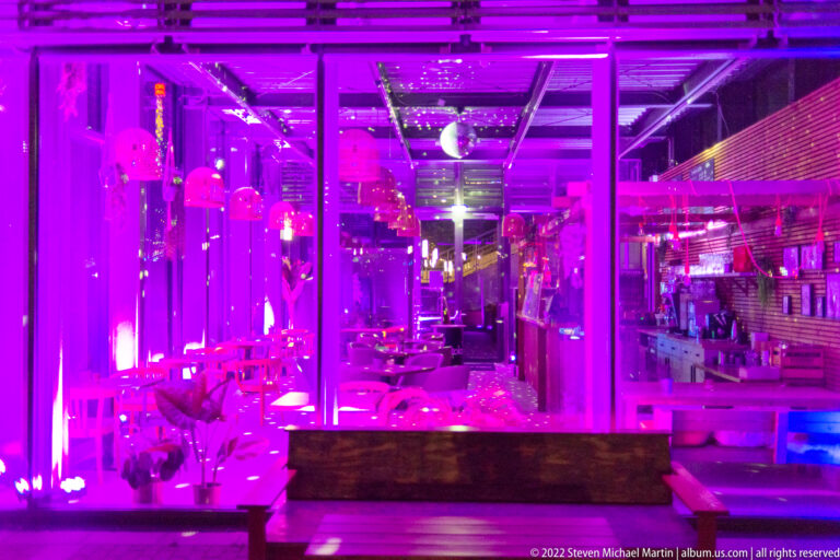 Contemporary bar or lounge interior glowing with magenta lighting, reflective glass, seating, and exposed structural elements.