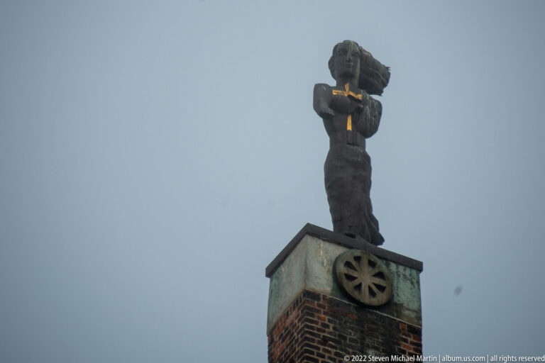 Weathered dark figure on a high brick architectural base, holding a small gilded object against a gray sky.