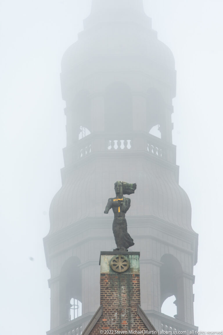 Small dark church sculpture with gilded detail aligned against the fog-obscured tower of St. Catherine’s Church.