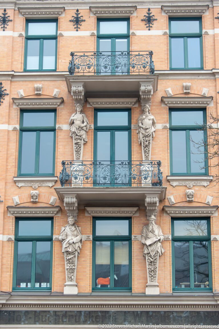 Ornate brick façade with green windows, wrought-iron balconies, and sculpted female figures between window bays.