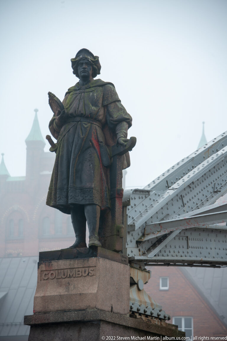 Bronze Christopher Columbus statue on a pedestal labeled “COLUMBUS,” with riveted bridgework and foggy buildings behind.
