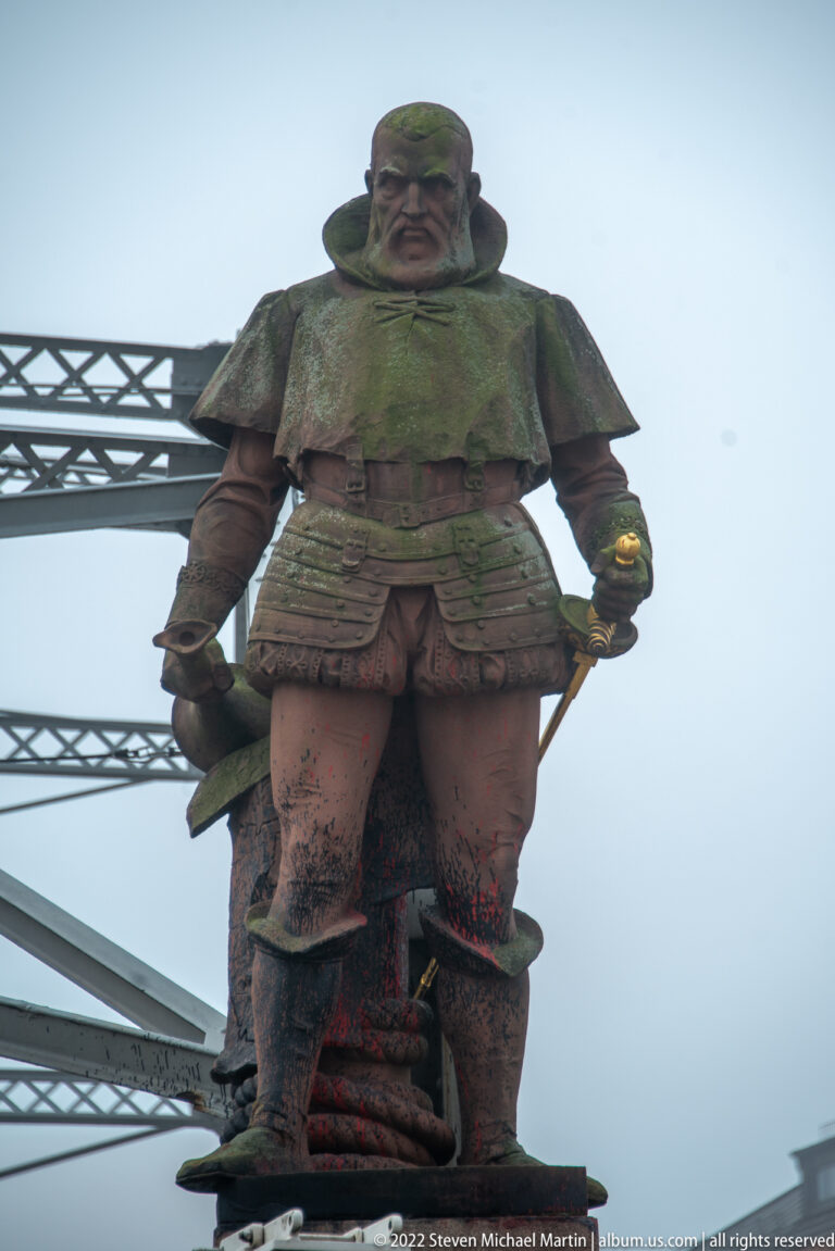 Bronze explorer statue in historical dress beside riveted steel bridgework against a fog-white sky.