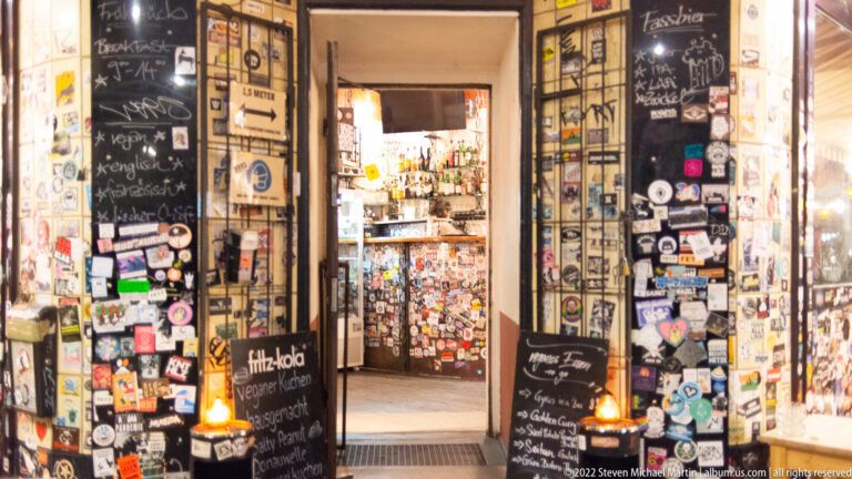 Sticker-covered doorway to a small bar or pub, with chalkboards, posters, and a brightly lit interior beyond.