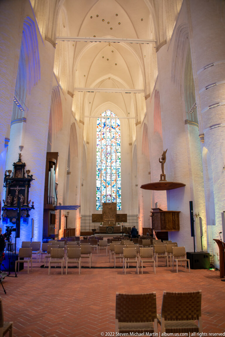 White brick nave of St. Catherine’s Church with pointed arches, modern seating, pulpit, and tall stained-glass window.