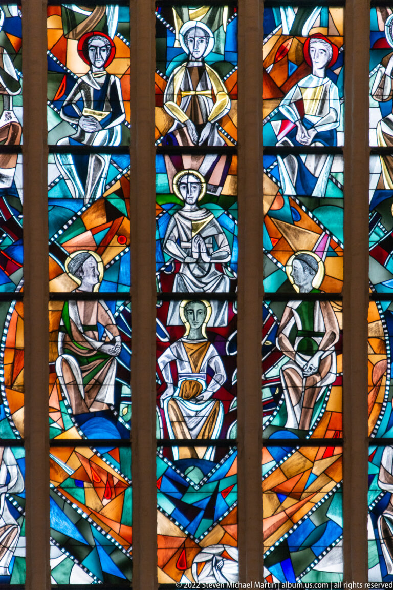 Close stained-glass detail with haloed figures in orange, blue, green, and turquoise panels divided by stone mullions.