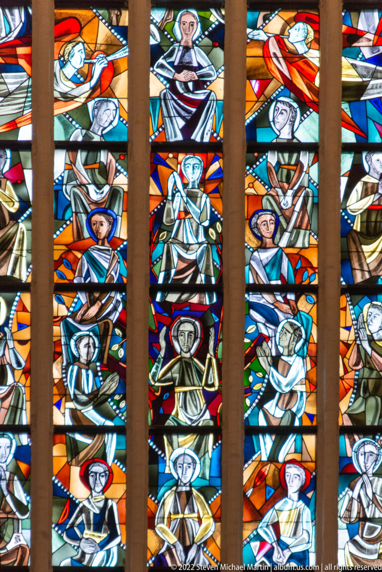 Stained-glass panel filled with rows of stylized haloed and robed figures in vivid orange, blue, and green.
