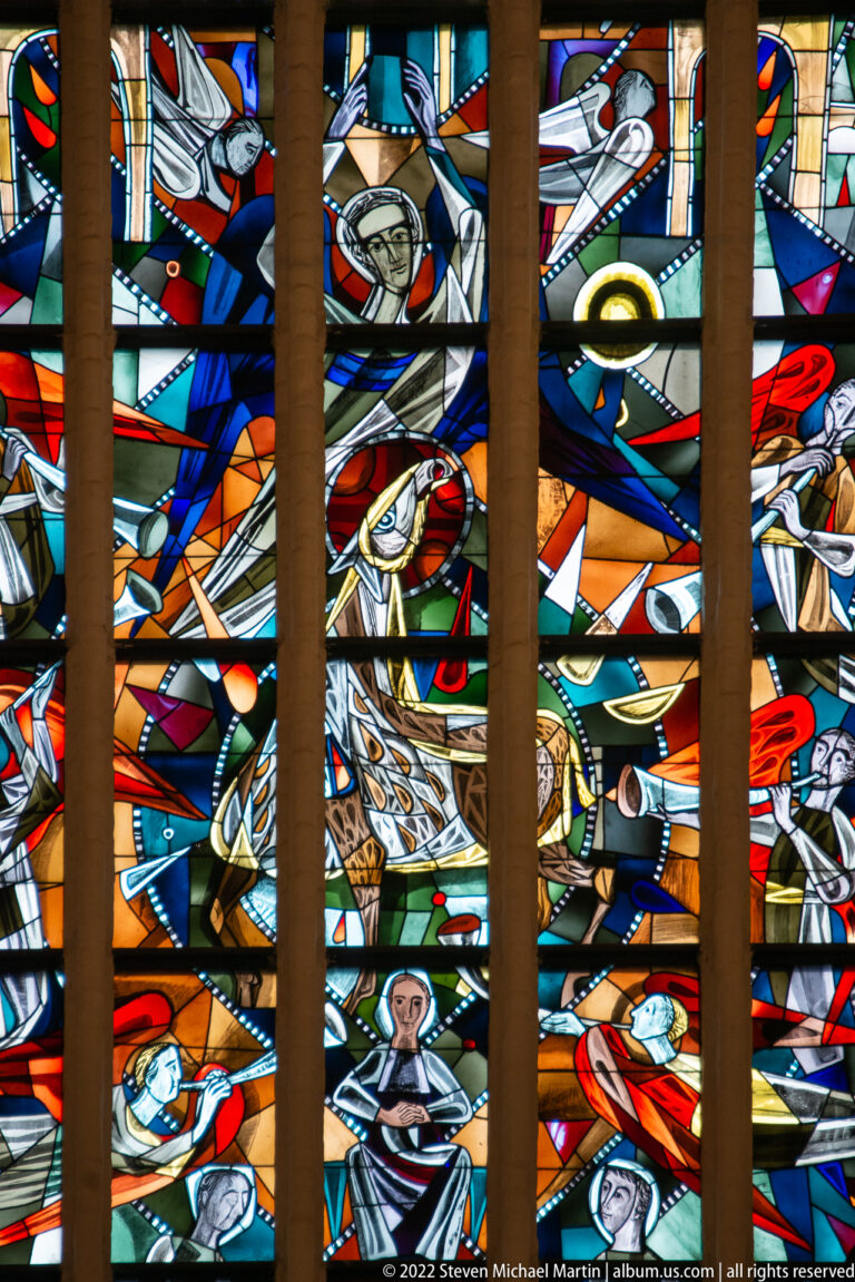 Vivid stained glass with kneeling, reclining, and standing figures arranged among red, blue, green, and amber forms.