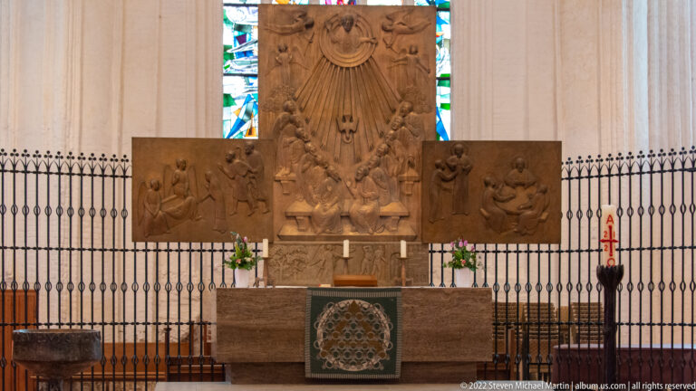 Sculptural church altar with central relief, side panels, candles, flowers, and black metal screen in front.