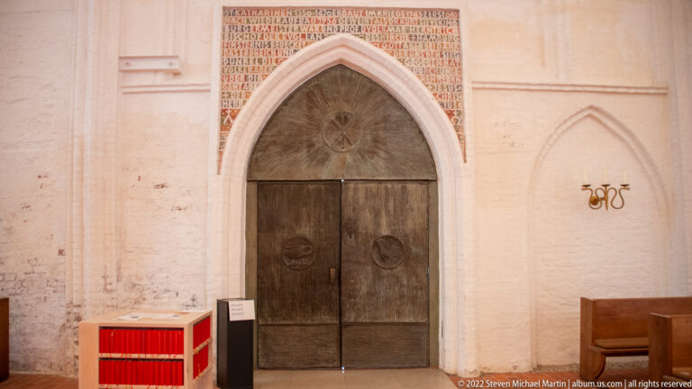 Pointed Gothic doorway with dark double wooden doors and inscription above in the pale brick church interior.