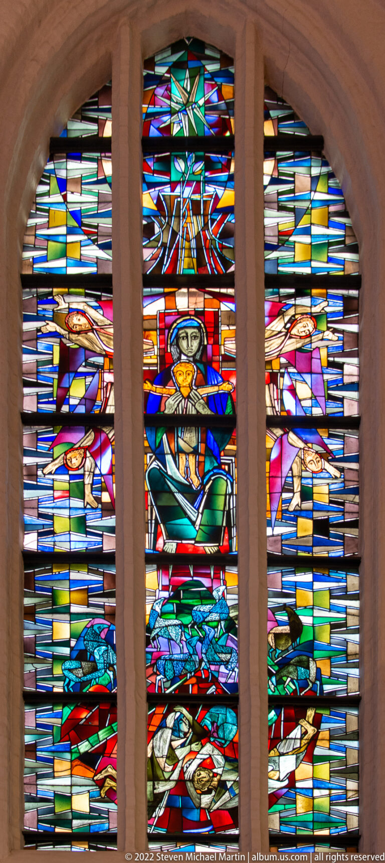 Three-light stained-glass window with central stylized figure and vivid horizontal bands of red, blue, yellow, and green.