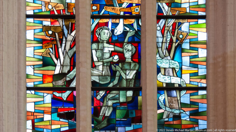 Modern stained-glass window with angular human figures and fragmented red, blue, yellow, and green geometric fields.