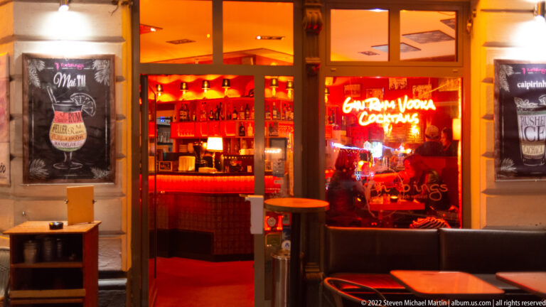Orange-red cocktail bar interior seen through a street window, with bottle shelves, neon sign, and outdoor tables.