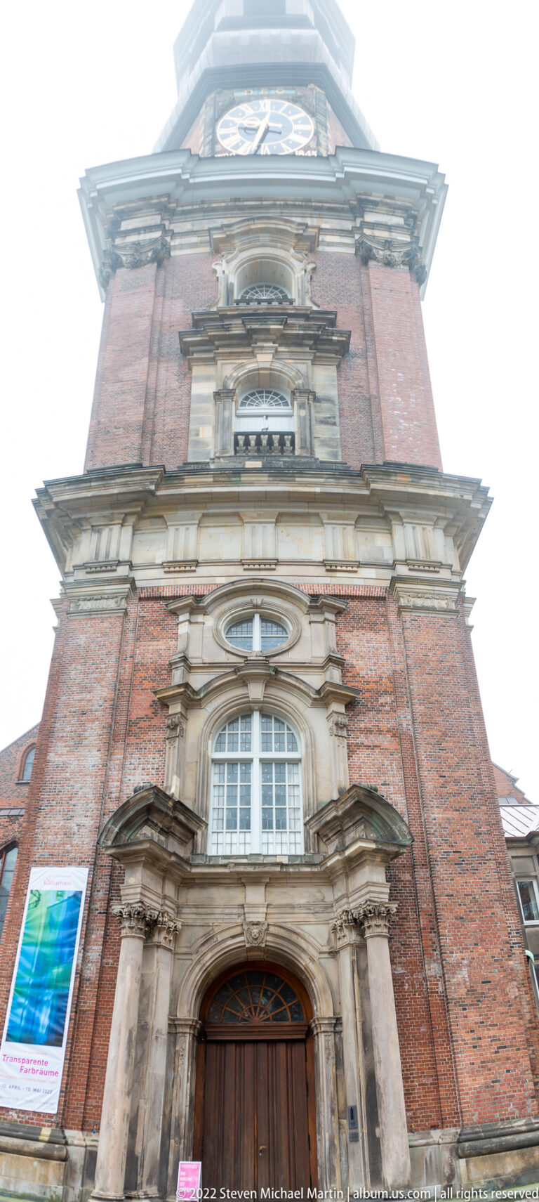 Ornate pale-stone church portal with curved pediments and wooden doors set into the red-brick tower of St. Catherine’s.