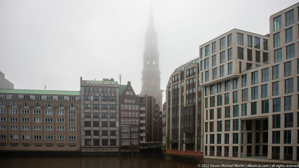 Hamburg canal lined by historic and modern buildings, with St. Catherine’s Church tower fading into fog in the distance.