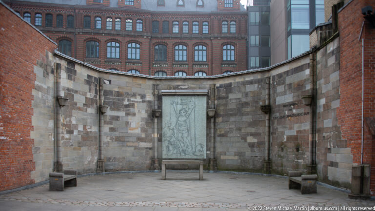 Freestanding memorial panel in a paved courtyard enclosed by a curved stone wall beside St. Nikolai ruins.