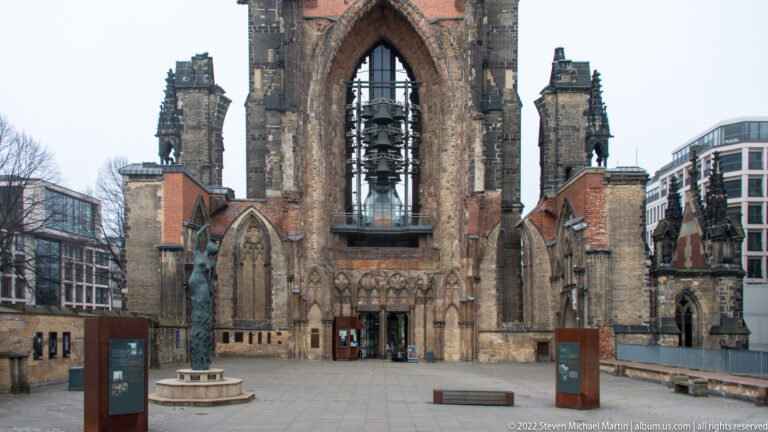 Open ruins of St. Nikolai with pointed arches, surviving tower, central sculpture, and modern buildings beyond.