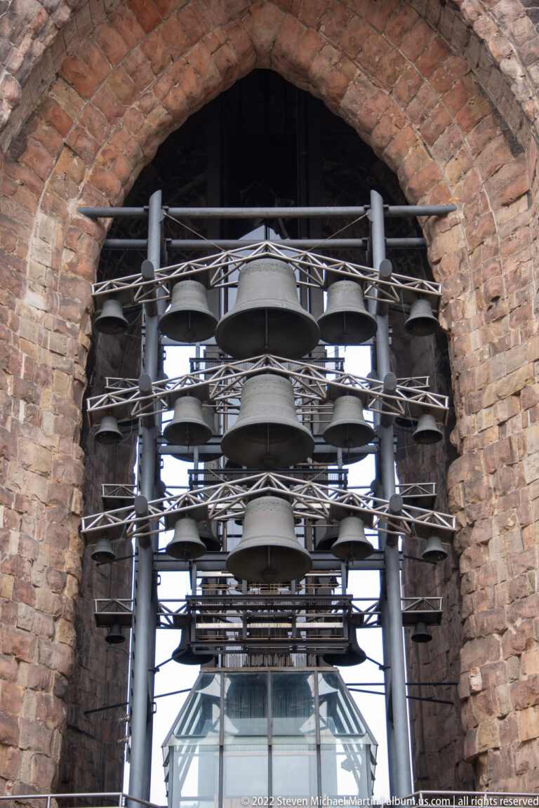 Multiple bells of different sizes hang on a modern metal framework within a tall weathered brick arch.