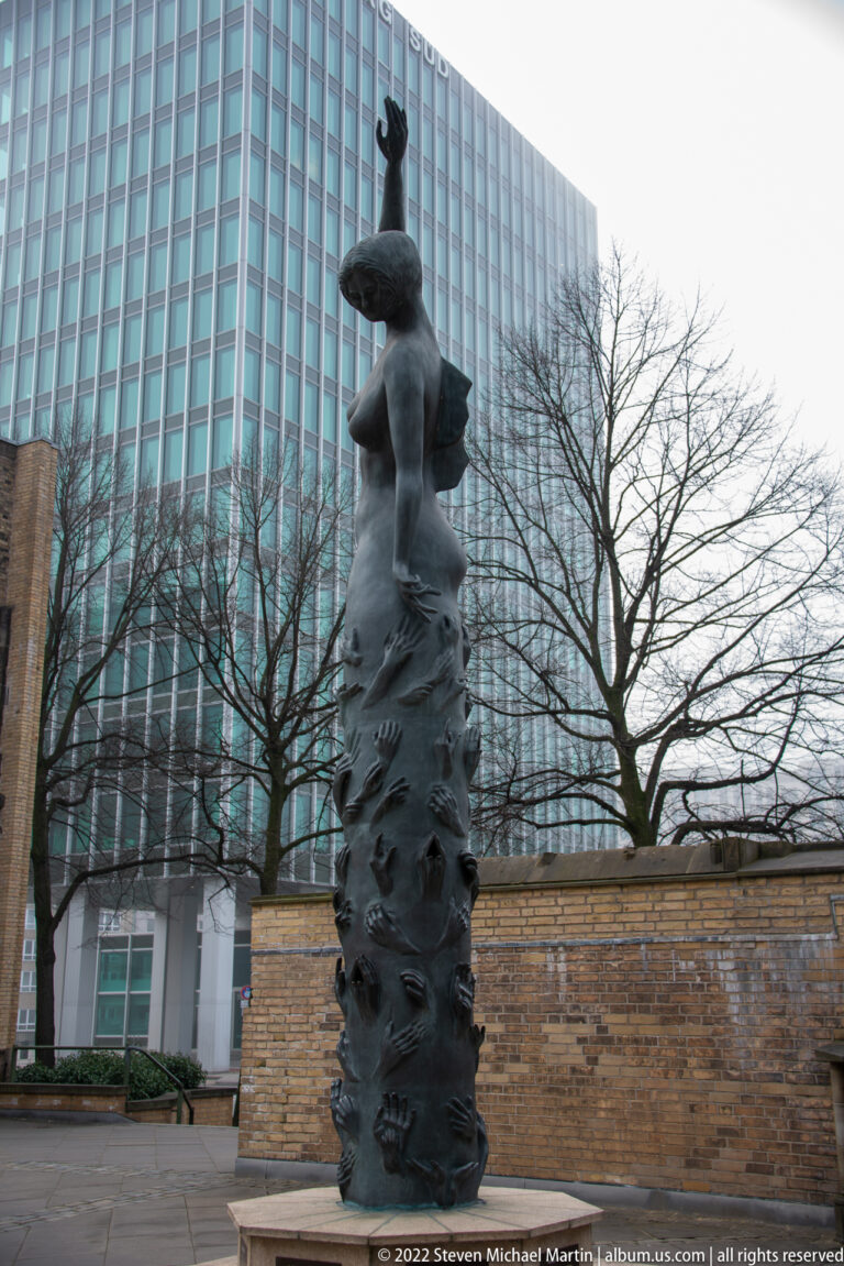 Tall dark bronze memorial sculpture with elongated human figure and textured column beside brick walls and modern buildings.