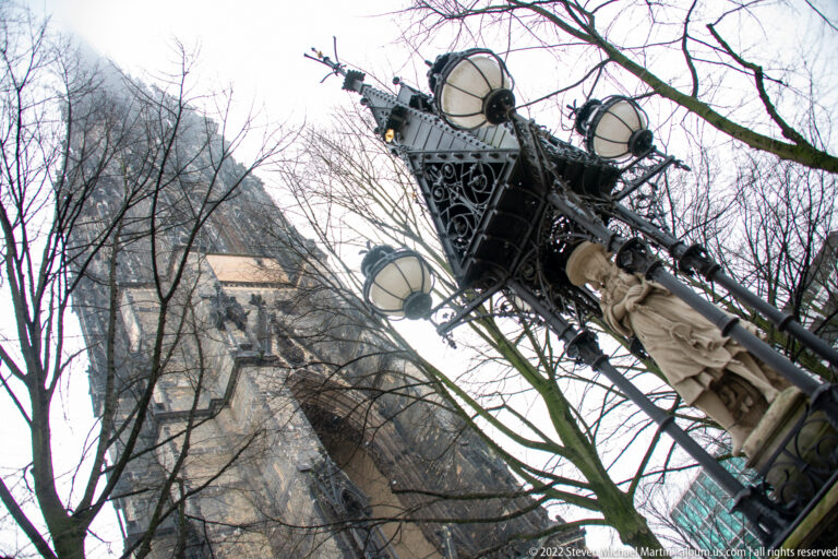 Ornate multi-globe streetlamp seen from below with bare branches and the St. Nikolai tower rising behind.