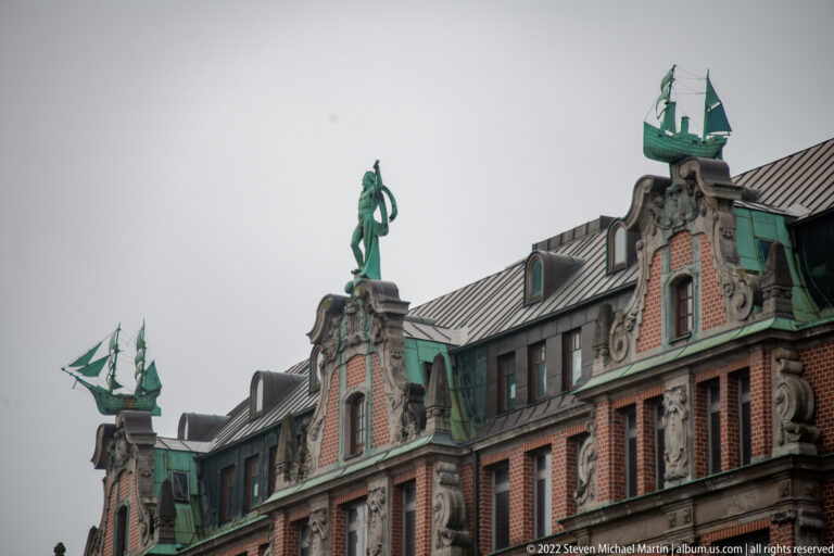 Three green-patinated sailing-ship sculptures crown ornate red-brick gables with carved stone figures and copper roof details.