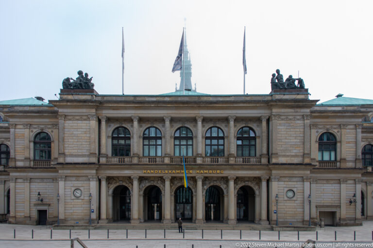 Symmetrical stone façade of Handelskammer Hamburg with arched entrances, tall windows, rooftop sculpture groups, and flags.