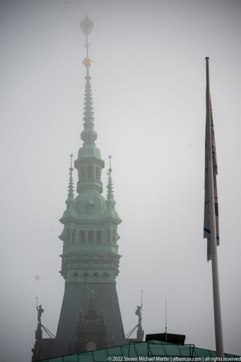 Green copper tower of Hamburg Rathaus rising into fog, with layered lanterns, pinnacles, and gilded summit ornament.