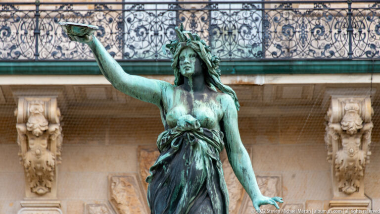 Green-patinated bronze female fountain figure with one arm extended, framed by ornate balcony ironwork and carved stone.