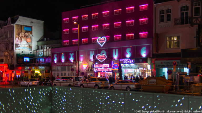 Night street in St. Pauli lined with pink neon venues, restaurants, cars, and illuminated pavement.