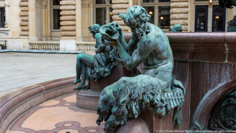 Two green-patinated seated bronze figures on the reddish stone rim of the Hygieia Fountain.