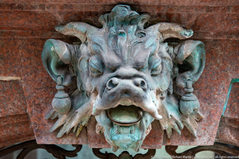 Close-up of a horned bronze grotesque mask with open waterspout mouth on the Hygieia Fountain.
