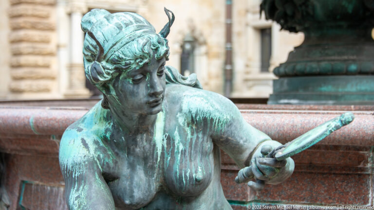 Green-patinated seated female bronze figure holding a long flat object on the Hygieia Fountain.