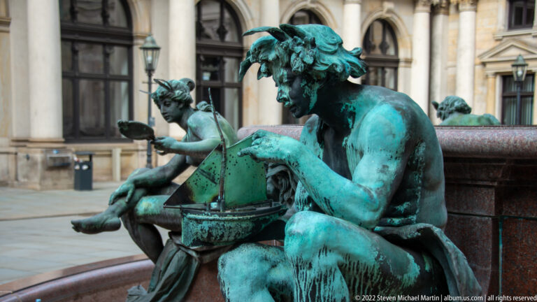 Green-patinated seated male bronze figure leaning over a rounded vessel on the Hygieia Fountain.