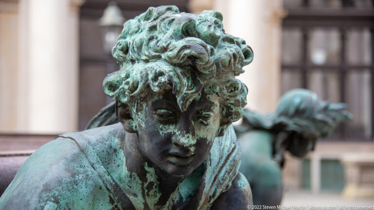 Close portrait of a weathered green bronze fountain figure with curled hair and lowered eyes.