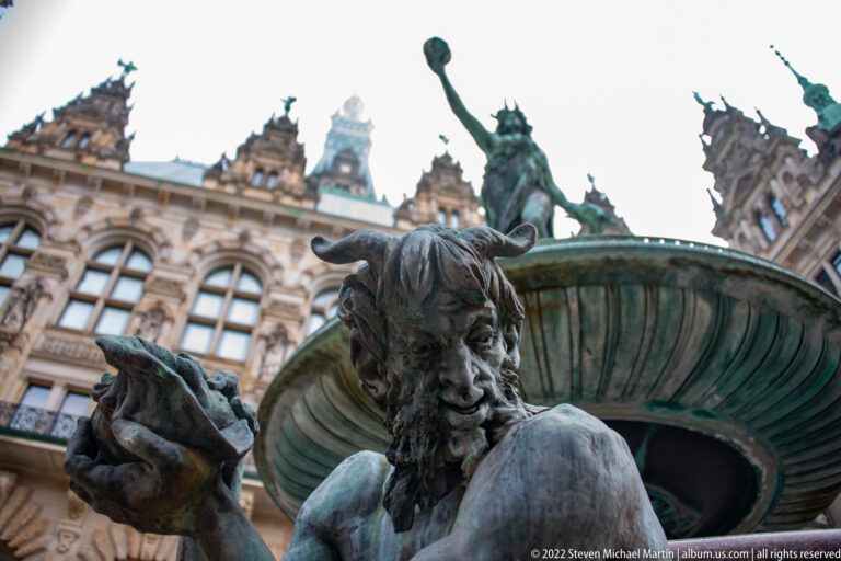 Bearded bronze figure beneath the broad central basin of the Hygieia Fountain, with Rathaus architecture behind.