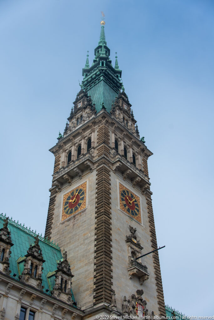 Low-angle view of Hamburg Rathaus clock tower with colorful clock faces, stone upper stages, and green copper roof.