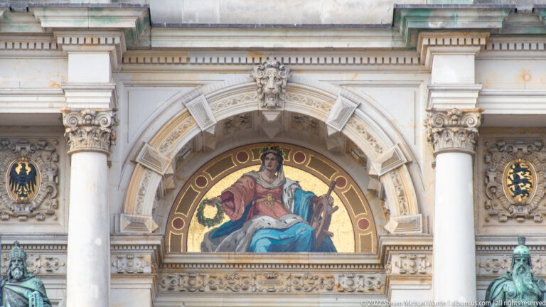 Semicircular mosaic of a seated robed woman above a Rathaus opening, flanked by columns and heraldic medallions.