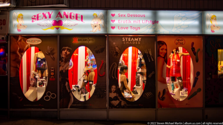Adult boutique storefront with oval display windows, lingerie mannequins, and signs for erotic goods and love toys.
