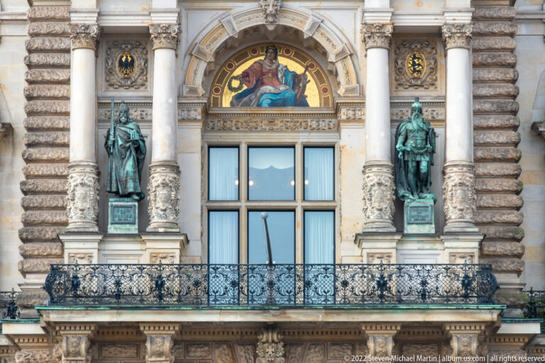 Ornate Hamburg Rathaus balcony with wrought iron, semicircular mosaic, and standing bronze statues on either side.
