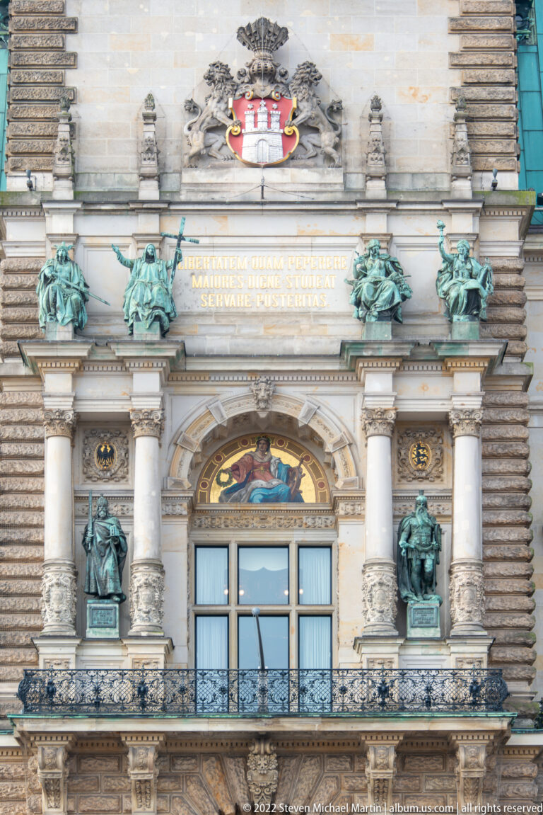 Symmetrical Hamburg Rathaus façade bay with balcony, mosaic, bronze figures, gilded inscription, and city coat of arms above.