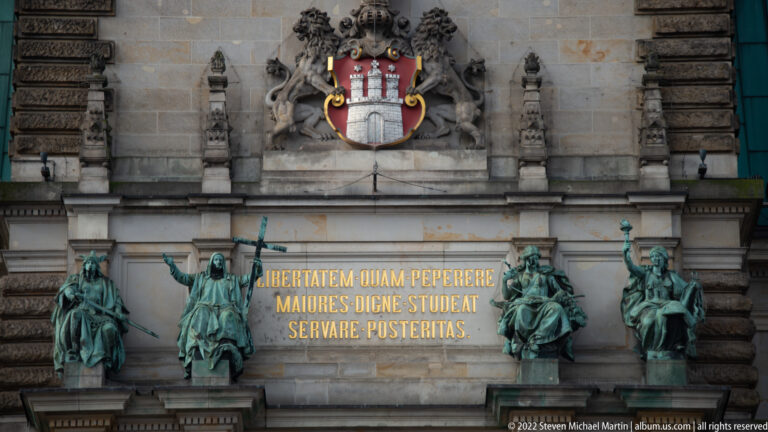 Hamburg coat of arms above a gilded Latin inscription and four bronze figures on an ornate Rathaus interior wall.
