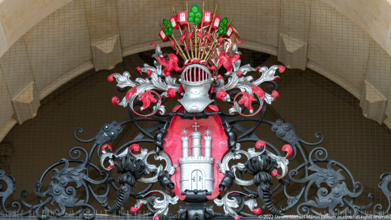 Ornate wrought-iron crest with Hamburg’s red-and-white coat of arms, plumed helmet, small banners, and black scrollwork.
