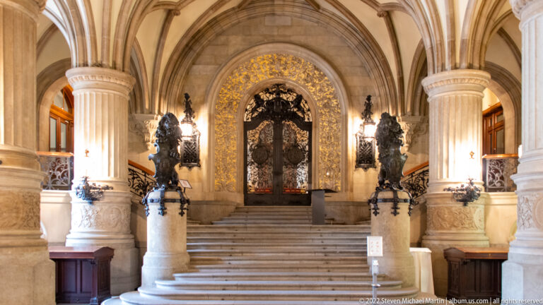 Stone ceremonial staircase inside Hamburg Rathaus leading to an ornate dark doorway framed by gilded decoration and sculptural lamps.